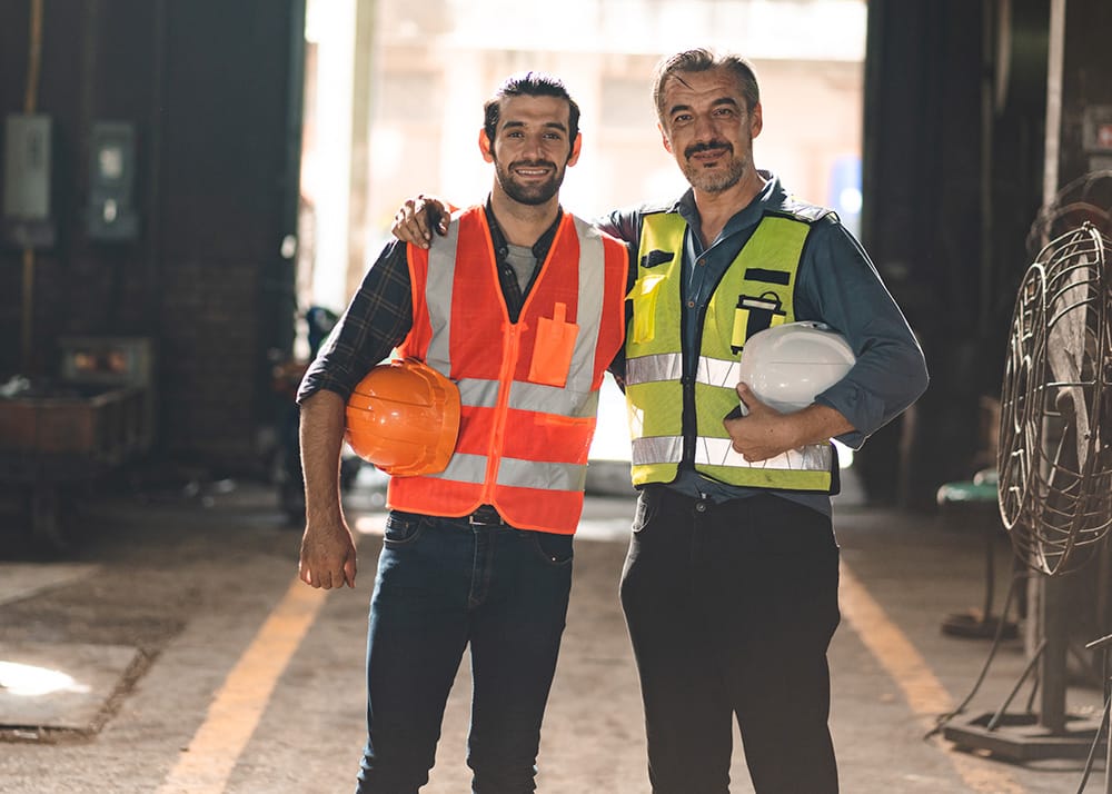 Portrait of senior and young male engineers and workers wearing safety vests and jacket while holding hardhat with arm around shoulder standing in front of machine in warehouse looking forward